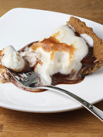 a piece of smores pie with a fork in it, on a white plate on a wood table