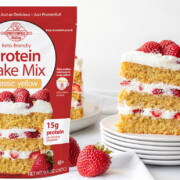 three layer cake of vanilla protei cake and whipped cream with fresh strawberries, next to a package of protein cake mix