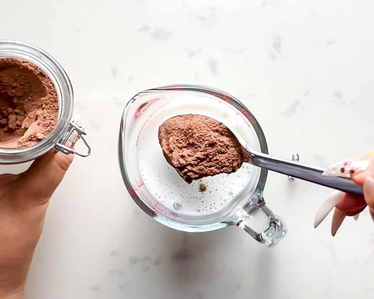 a spoonful of protein hot chocolate mix being added to a glass of warm milk