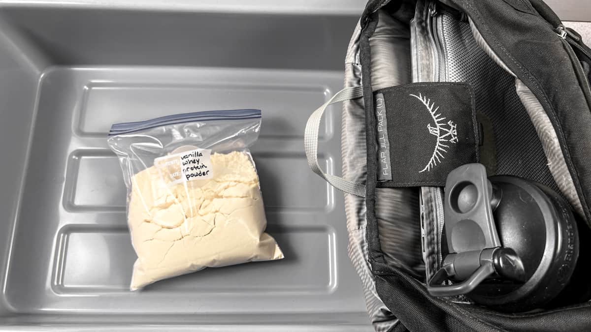 ziploc bag of vanilla protein powder in a grey TSA bin for airport security, next to a backpack with a shaker bottle inside.