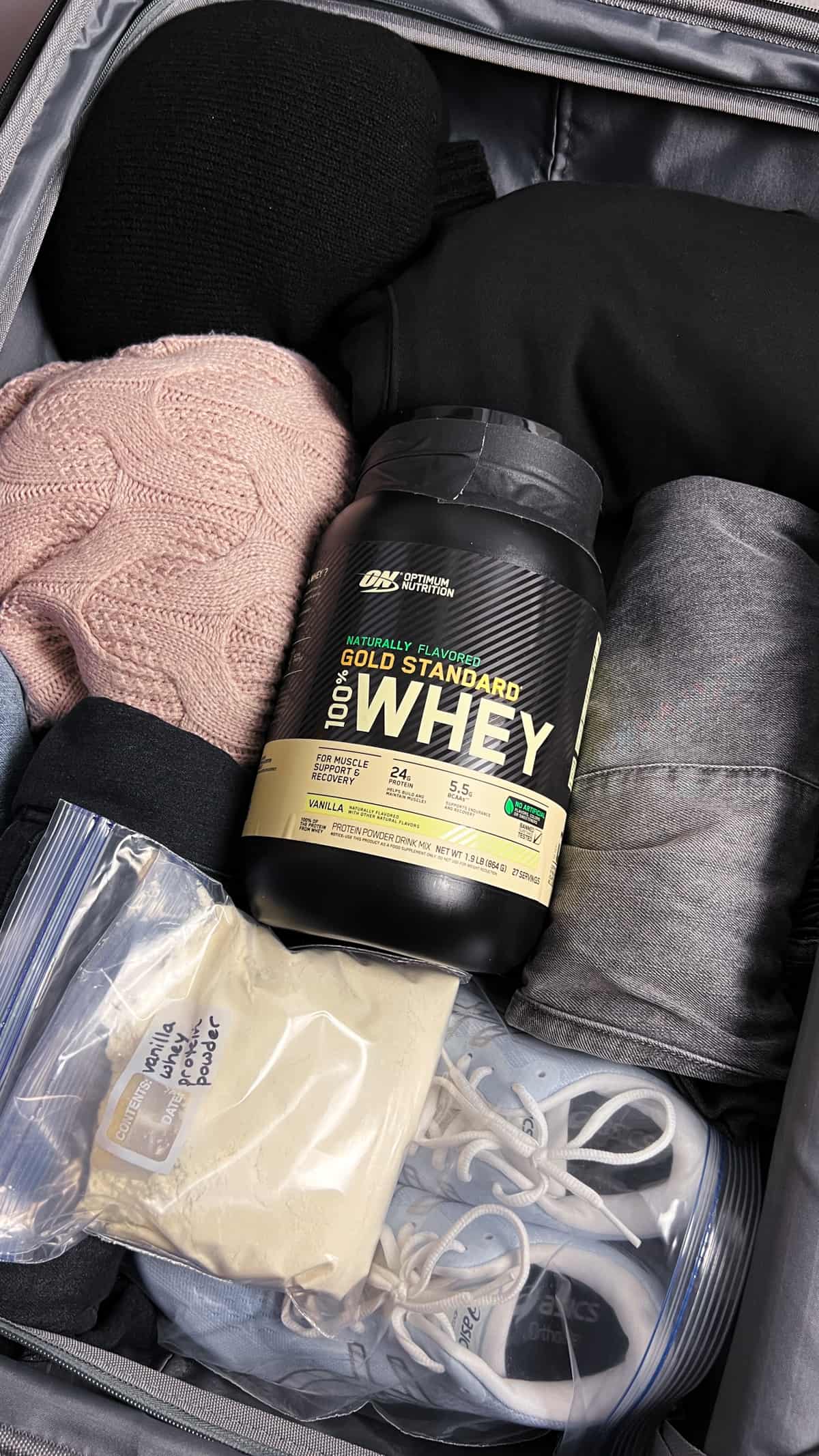 opened luggage showing clothing, sneakers, a bag of protein powder and a tub of protein powder.
