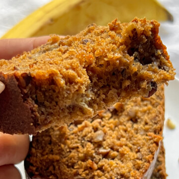 a sunlit bitten piece of protein banana bread being pulled off a stack, with a banana in the background.