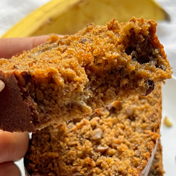 a sunlit bitten piece of protein banana bread being pulled off a stack, with a banana in the background.