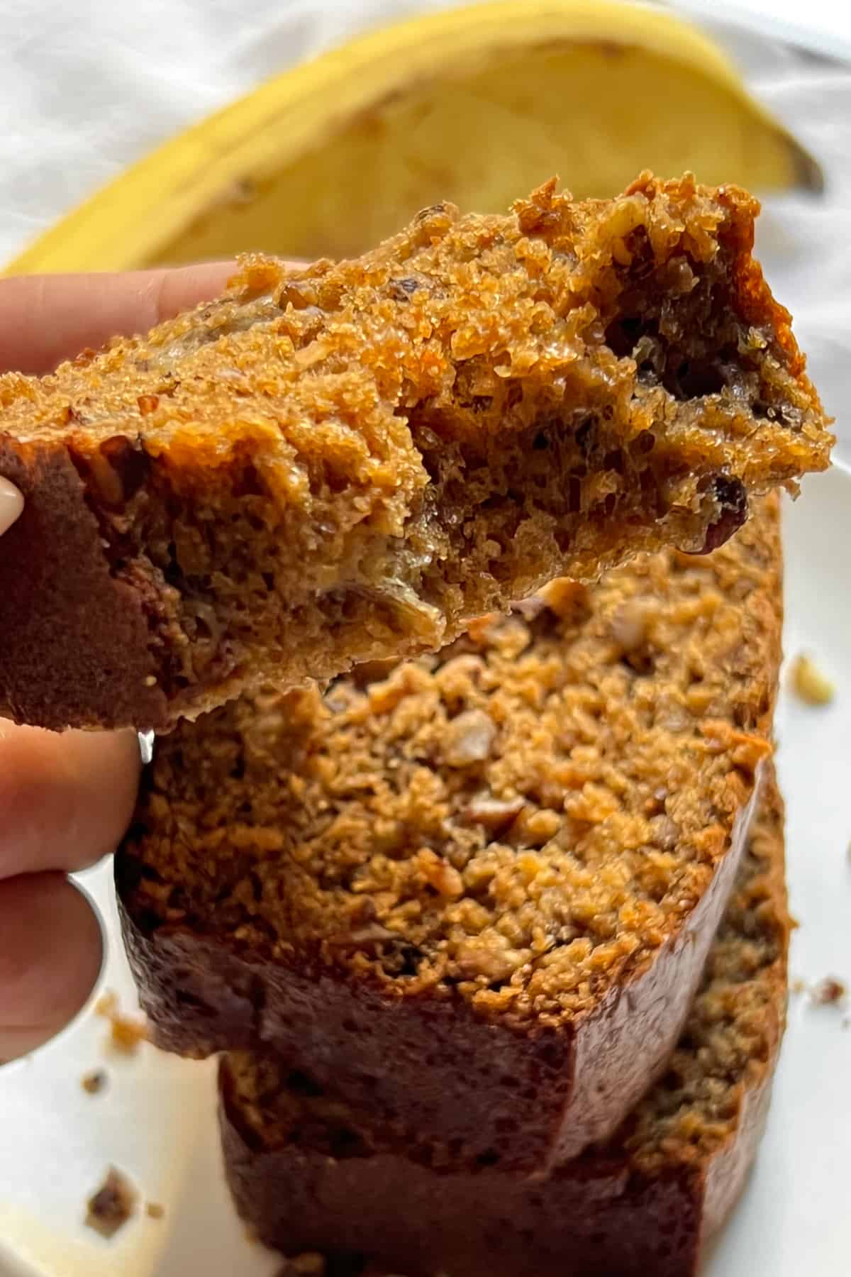 a sunlit bitten piece of protein banana bread being pulled off a stack, with a banana in the background.