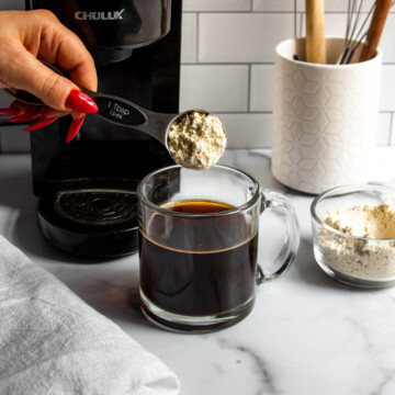 a spoonful of protein powder being added to hot black coffee in a glass mug