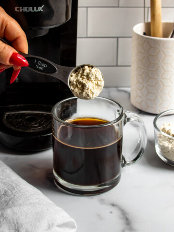 a spoonful of protein powder being added to hot black coffee in a glass mug