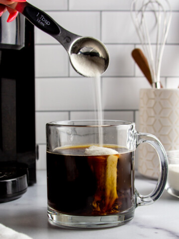 cup of black coffee in a glass mug with a tablespoon of collagen peptides being poured into it.