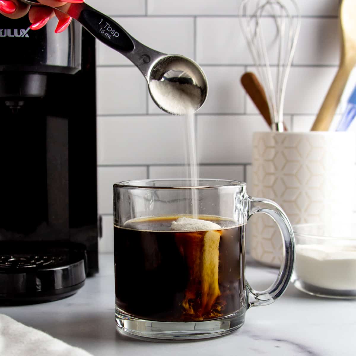 cup of black coffee in a glass mug with a tablespoon of collagen peptides being poured into it.