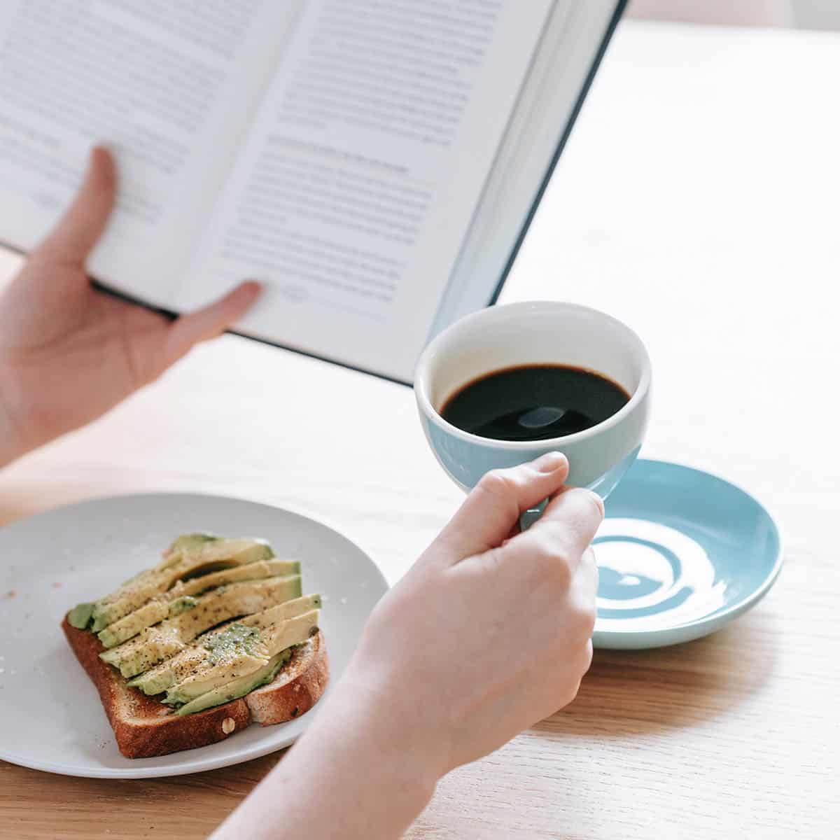 woman's hands with a book, coffee, and avocado toast.