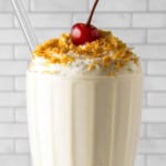 coconut protein shake in a milkshake glass with cream and a cherry on top.
