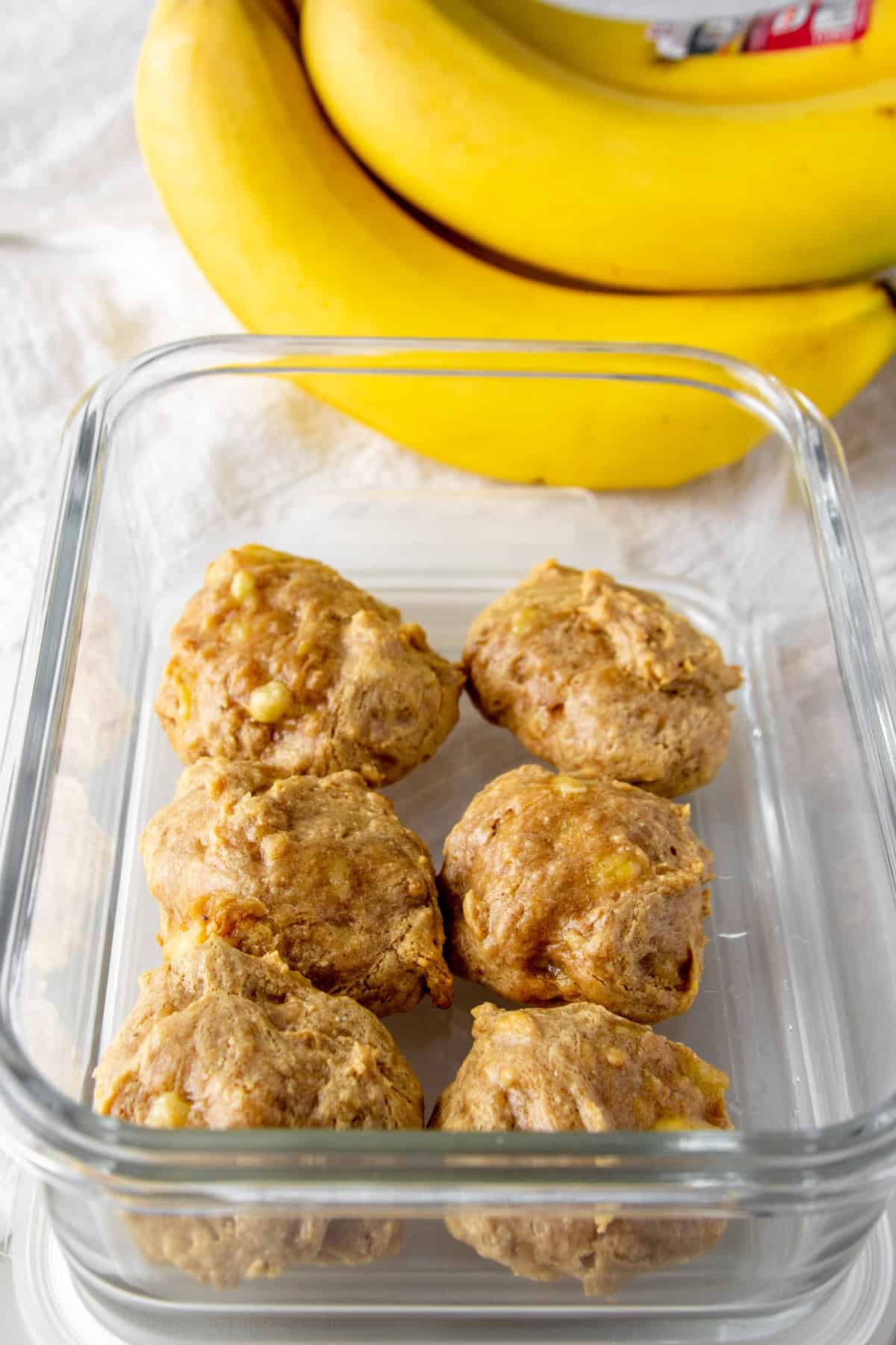 6 egg-free banana peanut butter protein cookies in a glass meal prep container in front of a bunch of bright yellow bananas.
