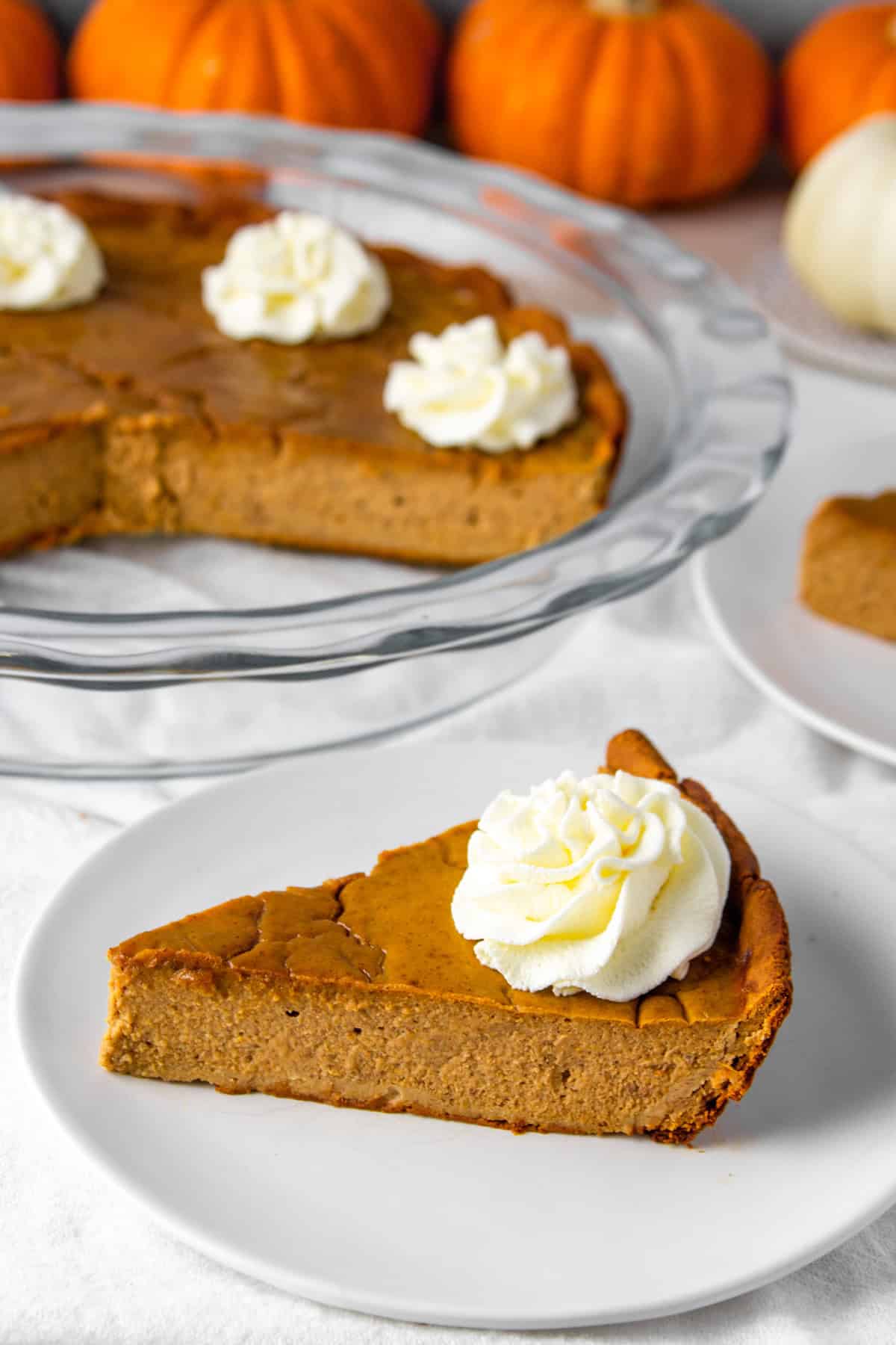 a slice of high protein pumpkin pie with sugar free whipped cream on top, with the whole pie and decorative mini pumpkins in the background.