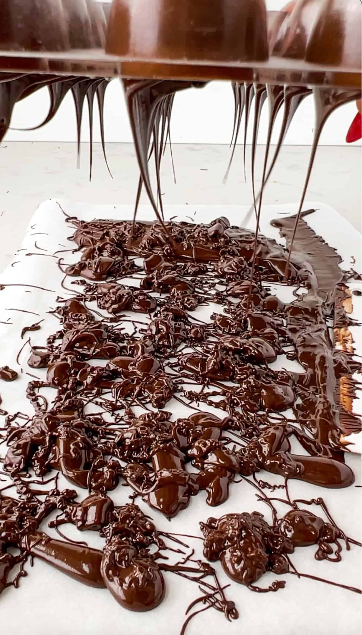 the chocolate mold being held upside down showing the excess melted chocolate dropping to parchment paper below.