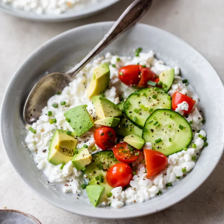 bowl of cottage cheese with cucumbers, tomato, and avocado, with a spoon.