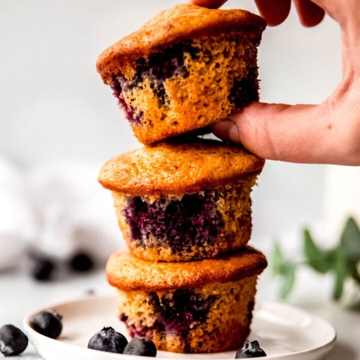 blueberry protein muffins stacked on a plate, one being picked up by hand.