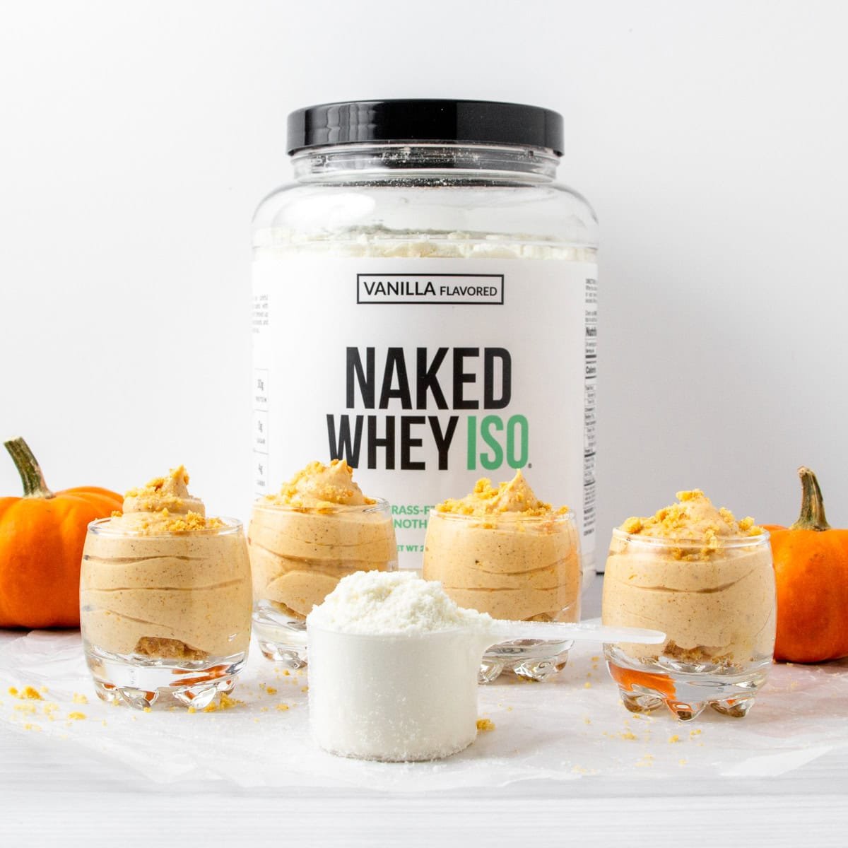 container of Naked Whey Iso with 4 servings of pumpkin cheesecake mousse, a scoop of protein powder, and two decorative mini pumpkins.